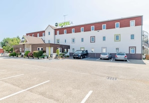Een hotel met een modern design, met een parkeerplaats met meerdere auto's, een klein terras en een bord met de naam van het hotel.