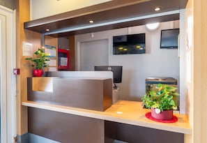 A reception desk with a computer, a potted plant, and a wall-mounted TV. - B&B Hôtel Lyon Saint-bonnet Mi-plaine (Saint-Bonnet-de-Mure)