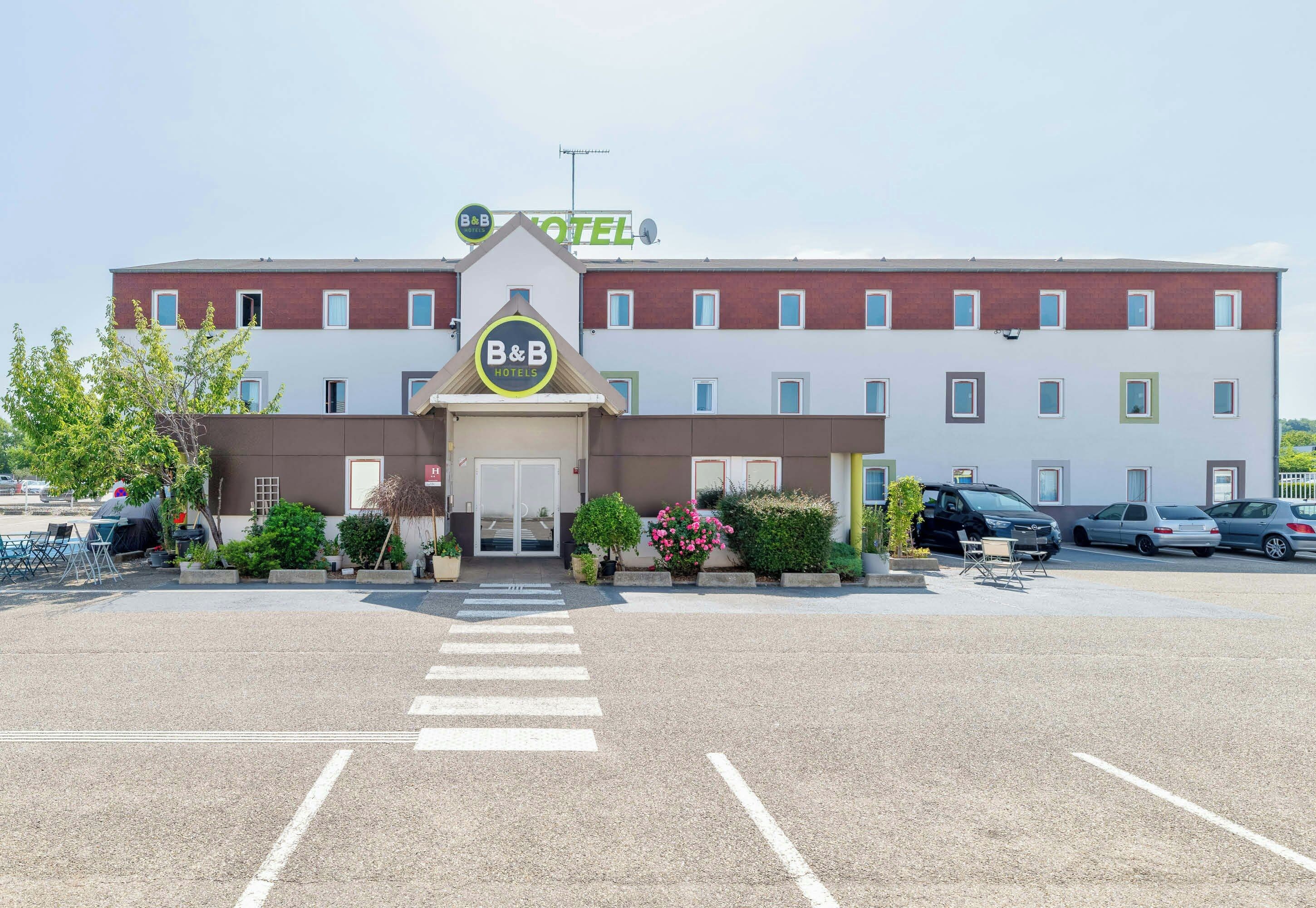 A B&B hotel with a central entrance, a parking lot, and a row of parked cars.