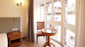 A room with a wooden nightstand, a chair, a round table, and large windows with a view of buildings. - Minotel Le Bourgogne (Evian-les-Bains)