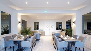 A hotel dining area with a central bar, multiple tables with chairs, and large windows. - Minotel Le Bourgogne (Evian-les-Bains)