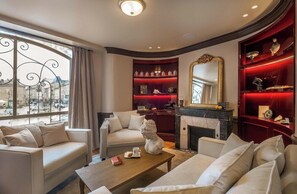 A spacious living room with a fireplace, a large window with curtains, and a red bookcase. - Hotel Des Prelats (Nancy)