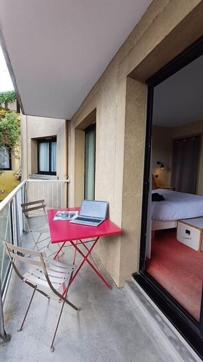 Un balcon avec une table rouge, une chaise et un ordinateur portable. La pièce adjacente contient un lit.