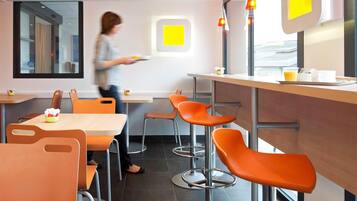 A modern dining area with high tables and orange chairs, a woman carrying a tray, and a window with a yellow sign.