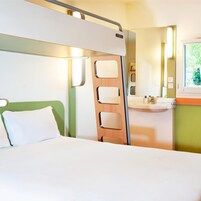 Una habitación pequeña con cama, un separador de madera, un lavamanos y una ventana.