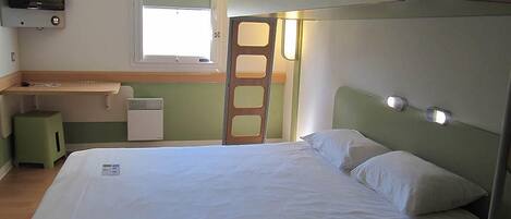A single bed with white linens, a small desk with a television, a window with blinds, and a green wall.