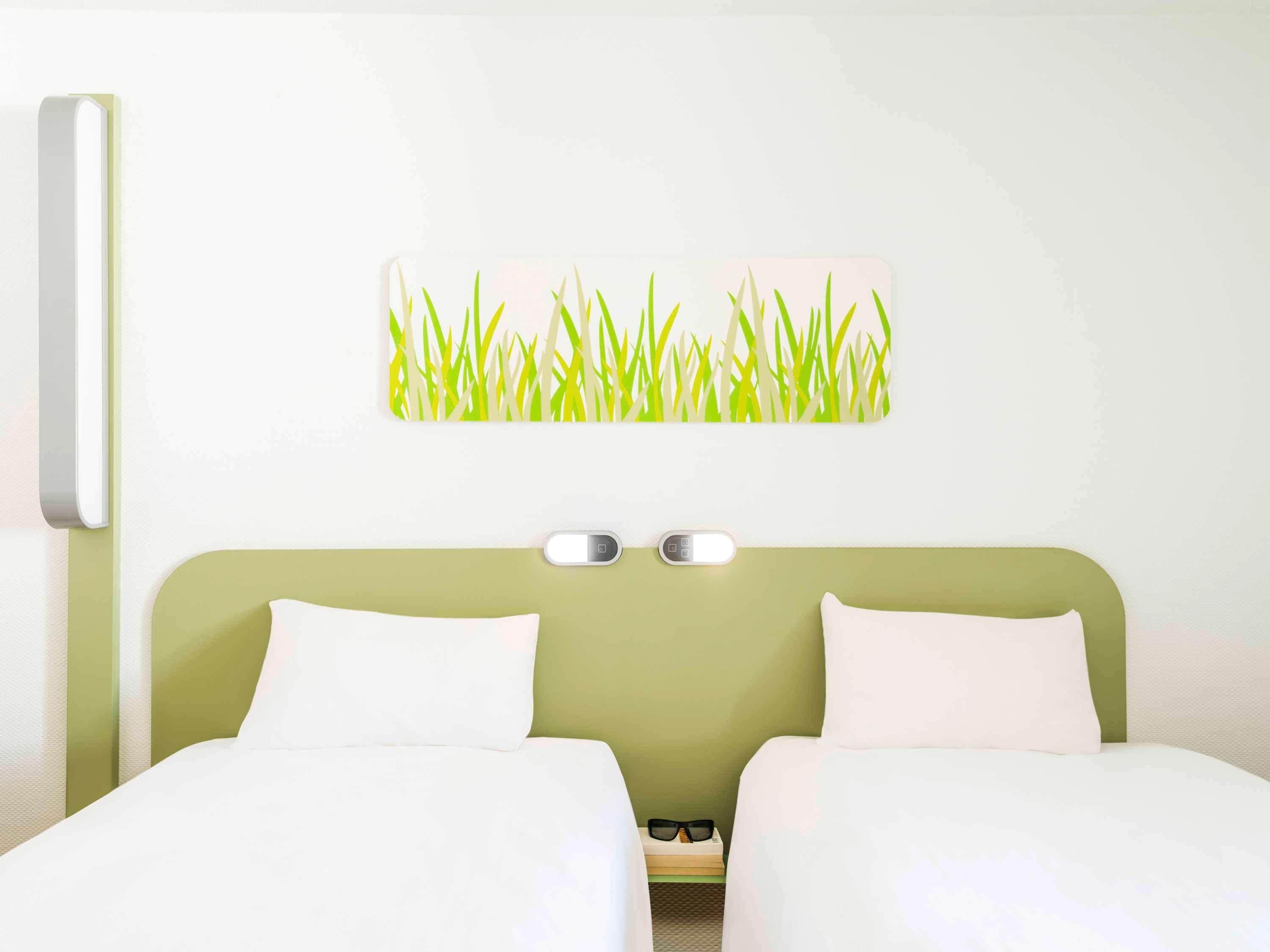 Une chambre d’hôtel avec deux lits, une tête de lit verte, une lampe fixée au mur et une décoration murale représentant de l’herbe.