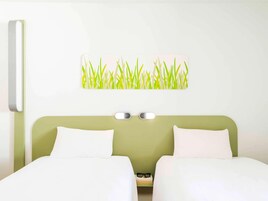 A hotel room with two beds, a green headboard, a wall-mounted light, and a wall art of grass.