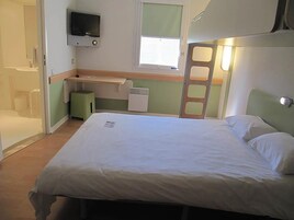 A single-bed room with a television mounted above the bed, a small desk with a chair, a window with blinds, and a door leading to another room.