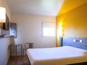 Una habitación de hotel con cama, una silla, una mesita, un espejo y una lámpara fijada a la pared.