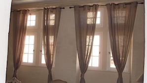 Ein Zimmer mit zwei Fenstern, jeweils mit Vorhängen, einem Sessel und einem kleinen Tisch mit einer Vase.
