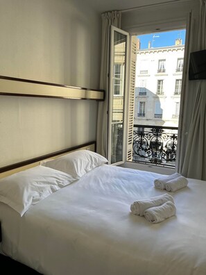 Een hotelkamer met een bed, twee kussens, een raam met gordijnen en uitzicht op een gebouw.