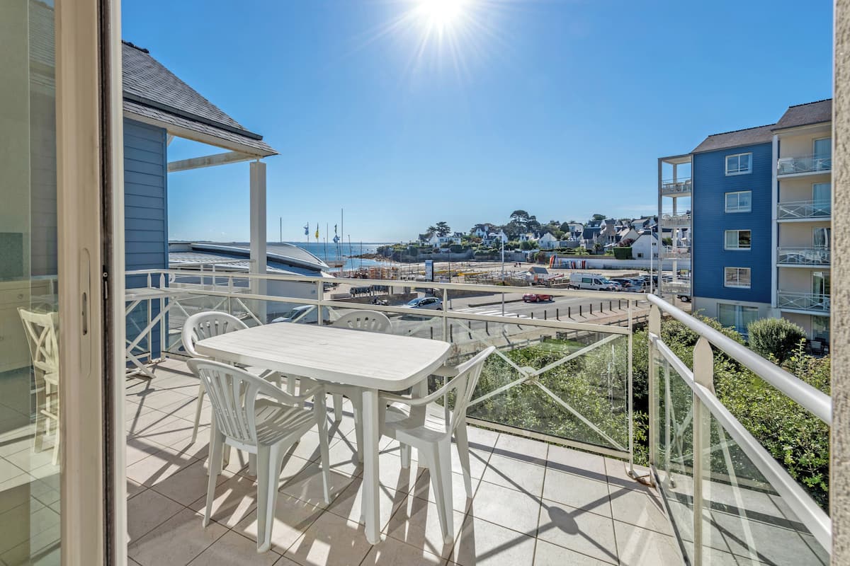 A balcony with a white table and chairs, overlooking a coastal town with buildings and a clear sky.
