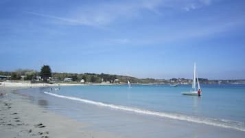 A beach with clear water, a sailboat, and a distant shoreline with buildings.
