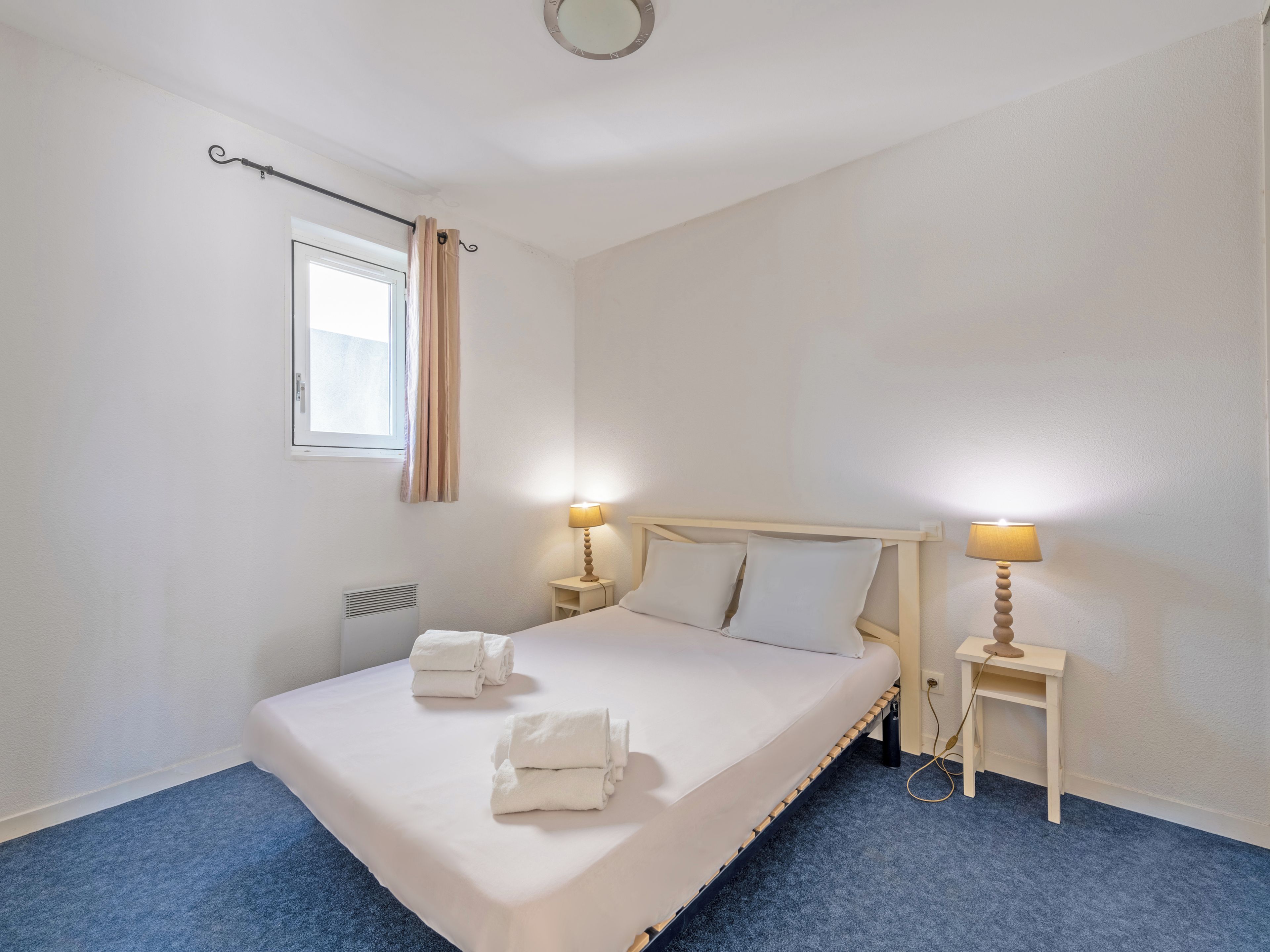 A neatly made bed with white linens and towels, two bedside tables with lamps, a window with curtains, and a radiator.