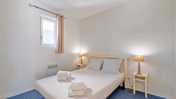 A neatly made bed with white linens and towels, two bedside tables with lamps, a window with curtains, and a radiator.