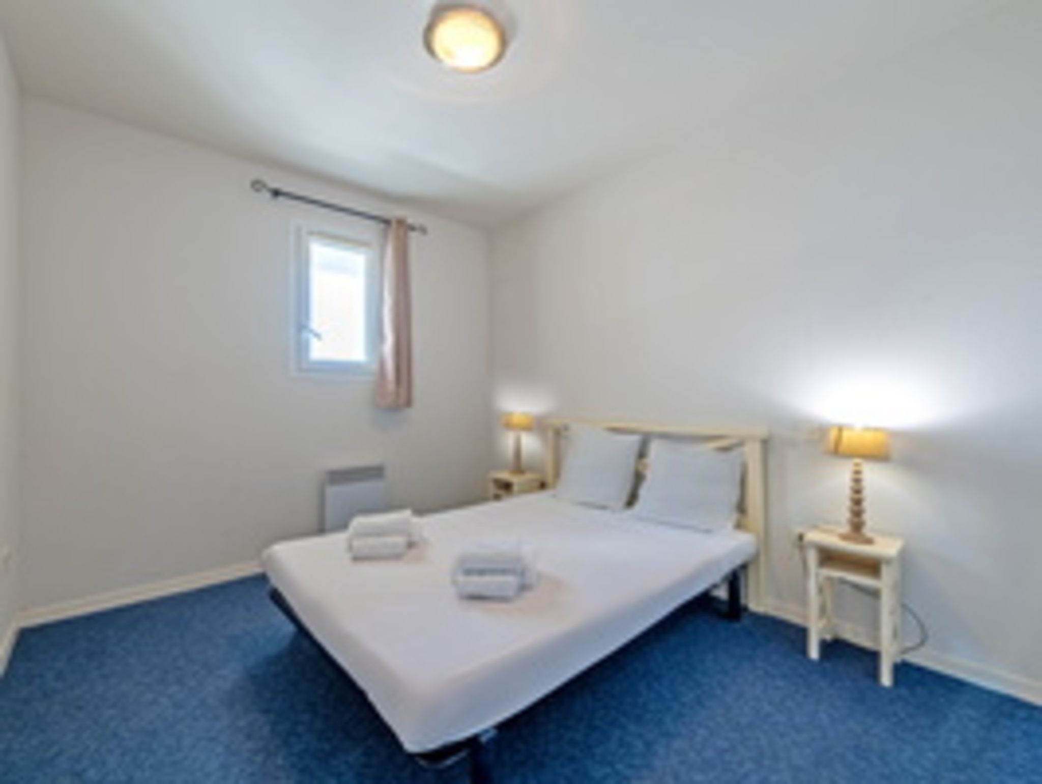 Habitación de hotel con una cama individual, dos mesitas de noche con lámparas, una ventana con cortinas y piso alfombrado.