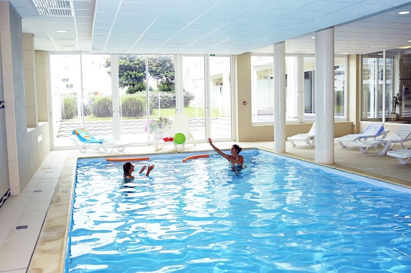 Ein Innenpool mit großen Glaswänden, gefliestem Boden und Liegestühlen rundherum.