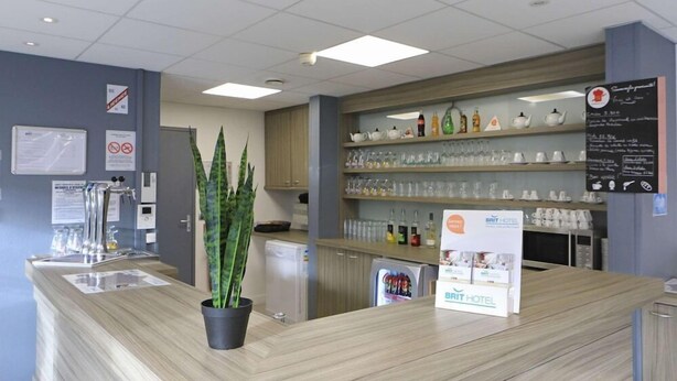 A hotel reception area with a wooden counter, a potted plant, a refrigerator, and shelves with glassware and bottles. - Brit Hotel Essentiel Dunkerque Gravelines (Gravelines)