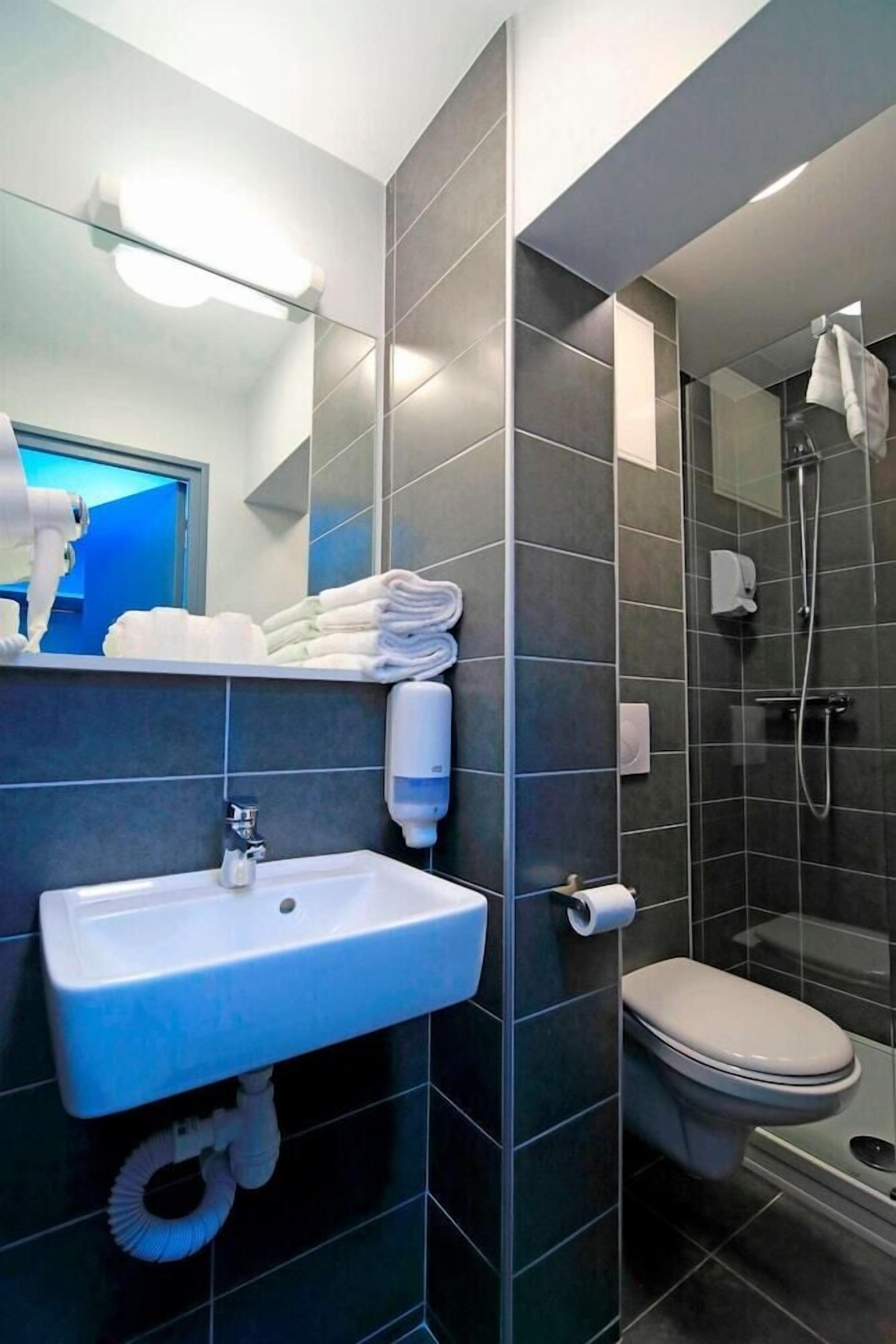 Une salle de bain moderne avec des carreaux foncés, un lavabo blanc, des toilettes et une douche séparée par une paroi de verre.
