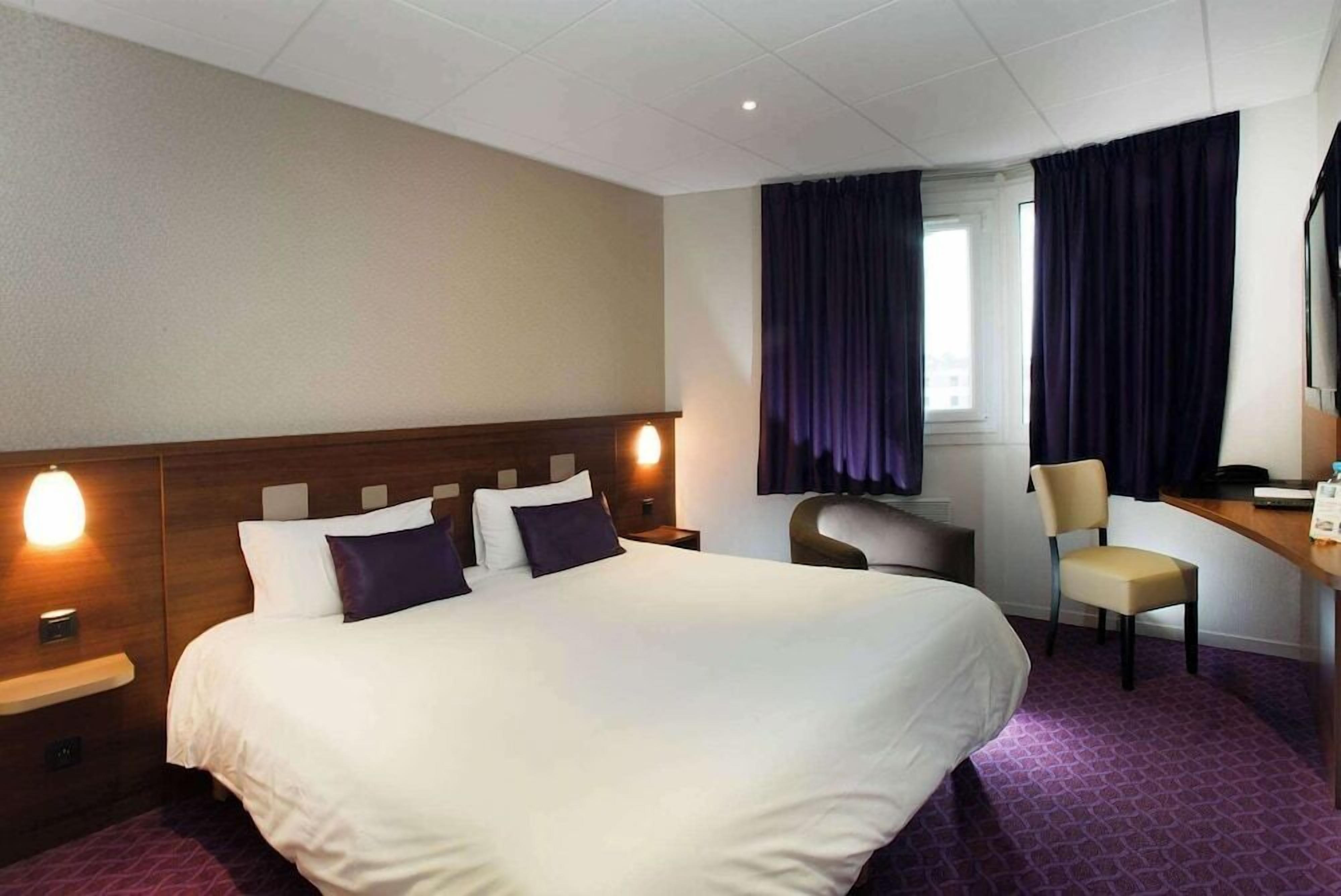 Una habitación de hotel con una cama grande, un escritorio con silla, una ventana con cortinas y una mesita de noche con lámpara.