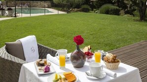 An outdoor breakfast setup with a wicker chair, a table with breakfast items, and a pool in the background. - Relais Du Silence Le Clos De Pradines (Saint-Quentin-la-Poterie)