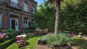 Un jardín con una mesa y sillas, un árbol grande y un edificio con balcón.