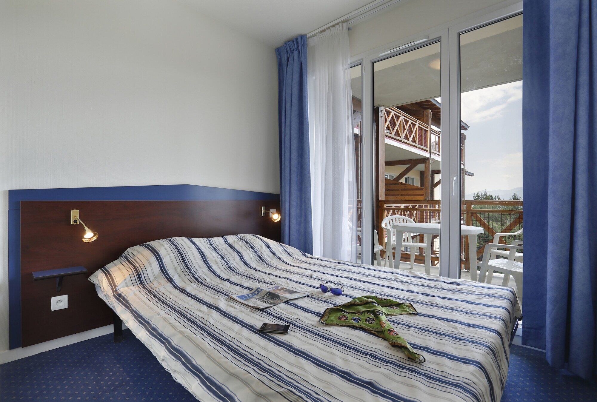 Ein Hotelzimmer mit Bett, Nachttisch, Lampe, einem Fenster mit Vorhängen und Blick auf den Außenbalkon.