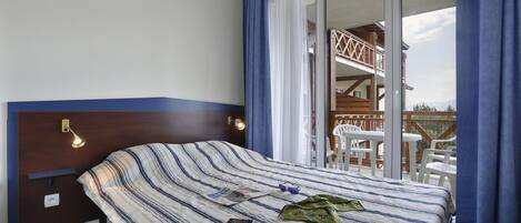 Ein Hotelzimmer mit Bett, Nachttisch, Lampe, einem Fenster mit Vorhängen und Blick auf den Außenbalkon.