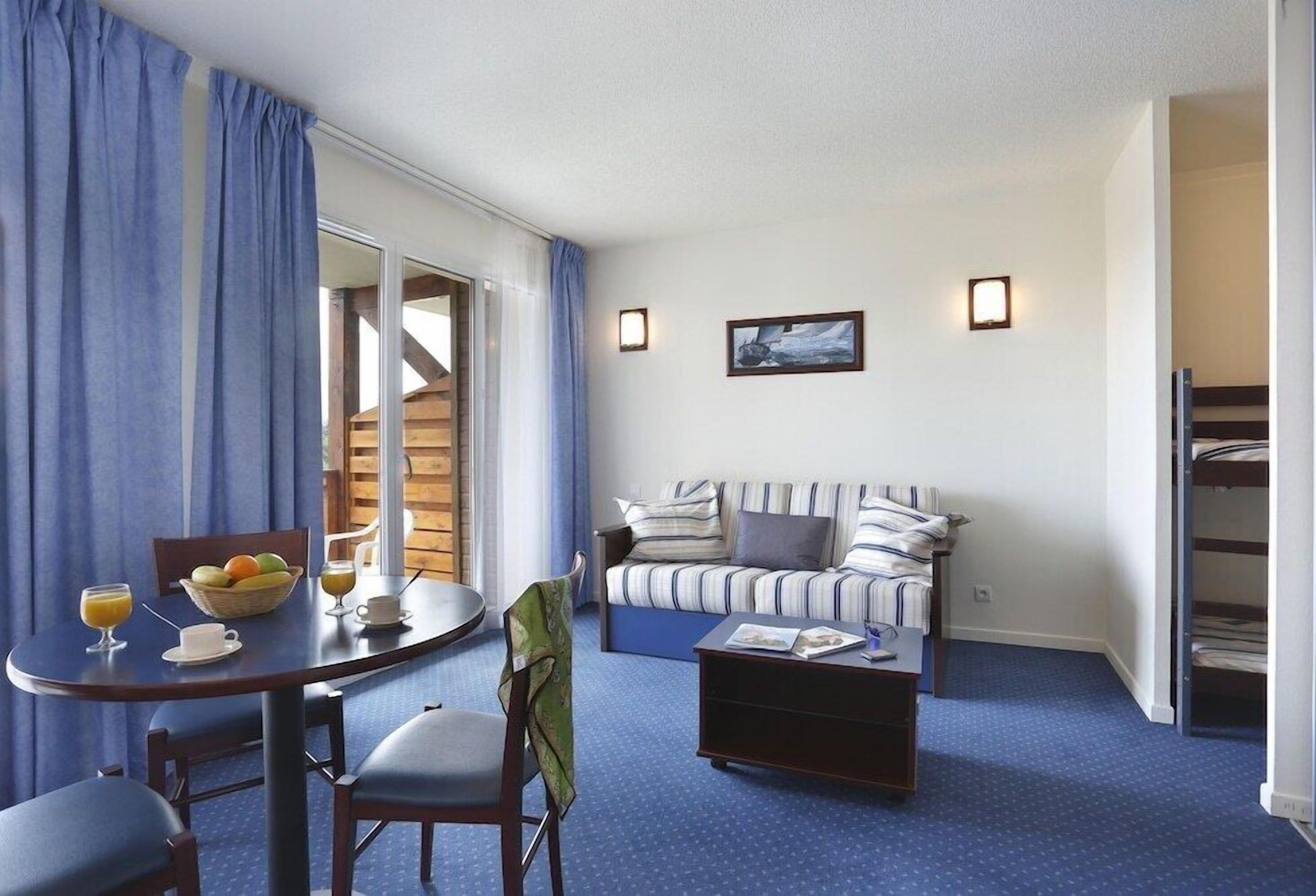 Een compacte hotelkamer met een bank, een klein tafeltje met een schaal fruit, een koffiekopje en een tijdschrift. Er is een raam met gordijnen, een wandlamp en een lijst met een afbeelding.