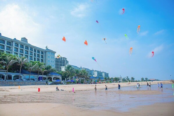 Ein Strand mit einem Hotel im Hintergrund, Palmen und Menschen im Sand.