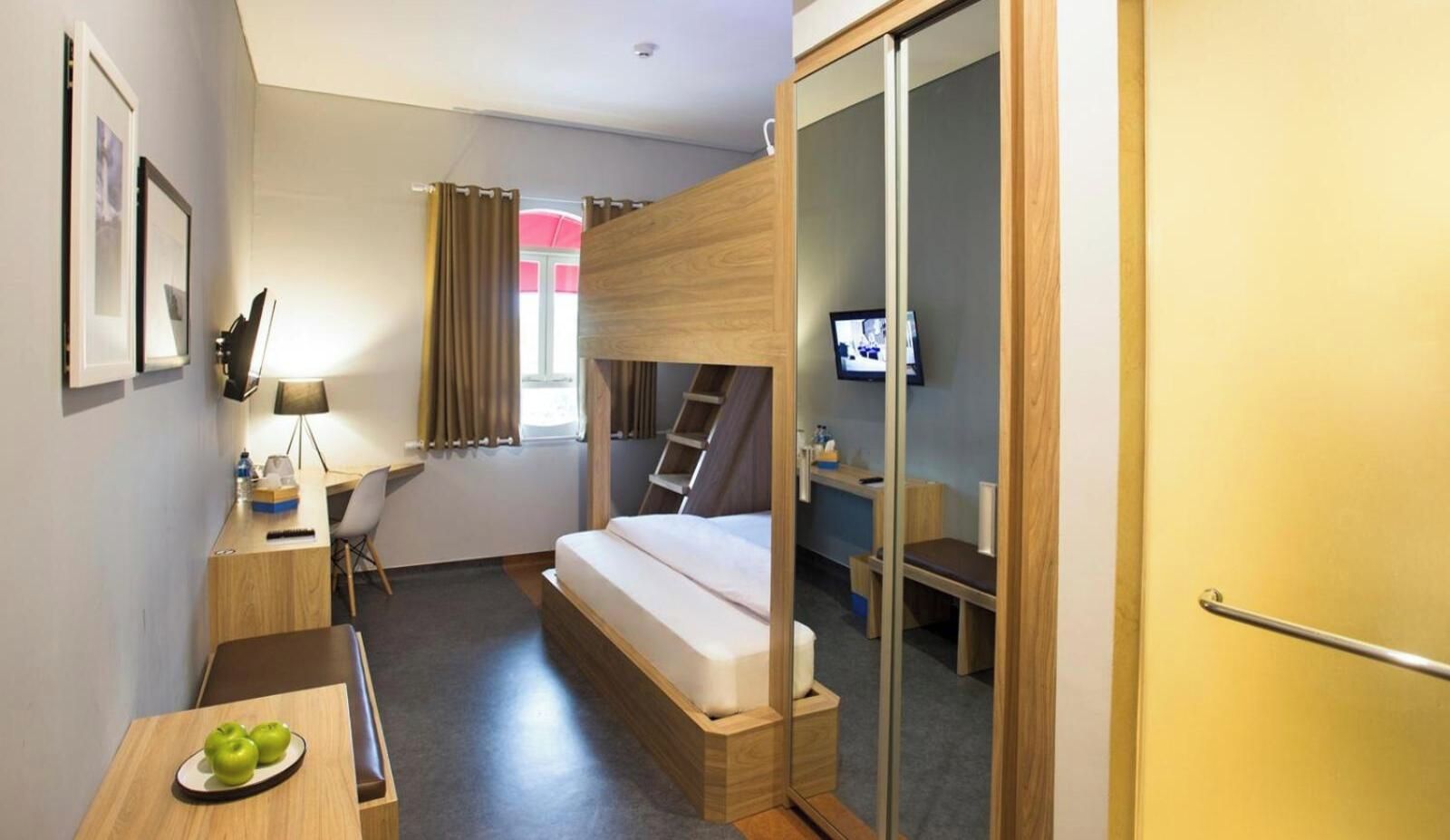 A compact hotel room with a bunk bed, a desk with a chair, a small table with apples, a television, and a sliding door.