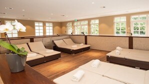 Ein Spa-Raum mit drei Massageliegen, Holzboden und großen Fenstern mit Grünpflanzen draußen.