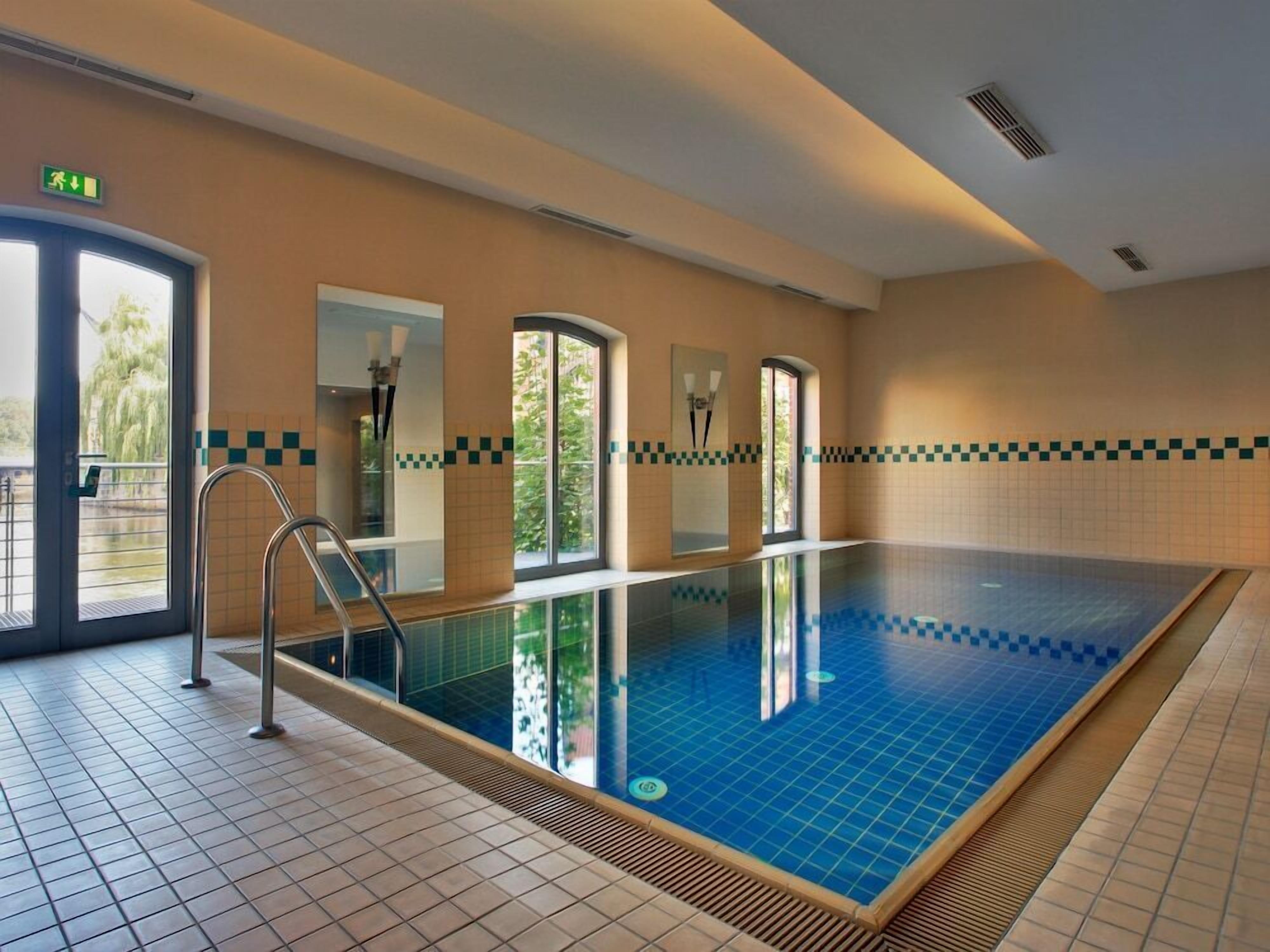 Ein großer, überdachter Pool mit Glaswänden, Fliesenboden und Blick auf die Grünanlagen draußen.