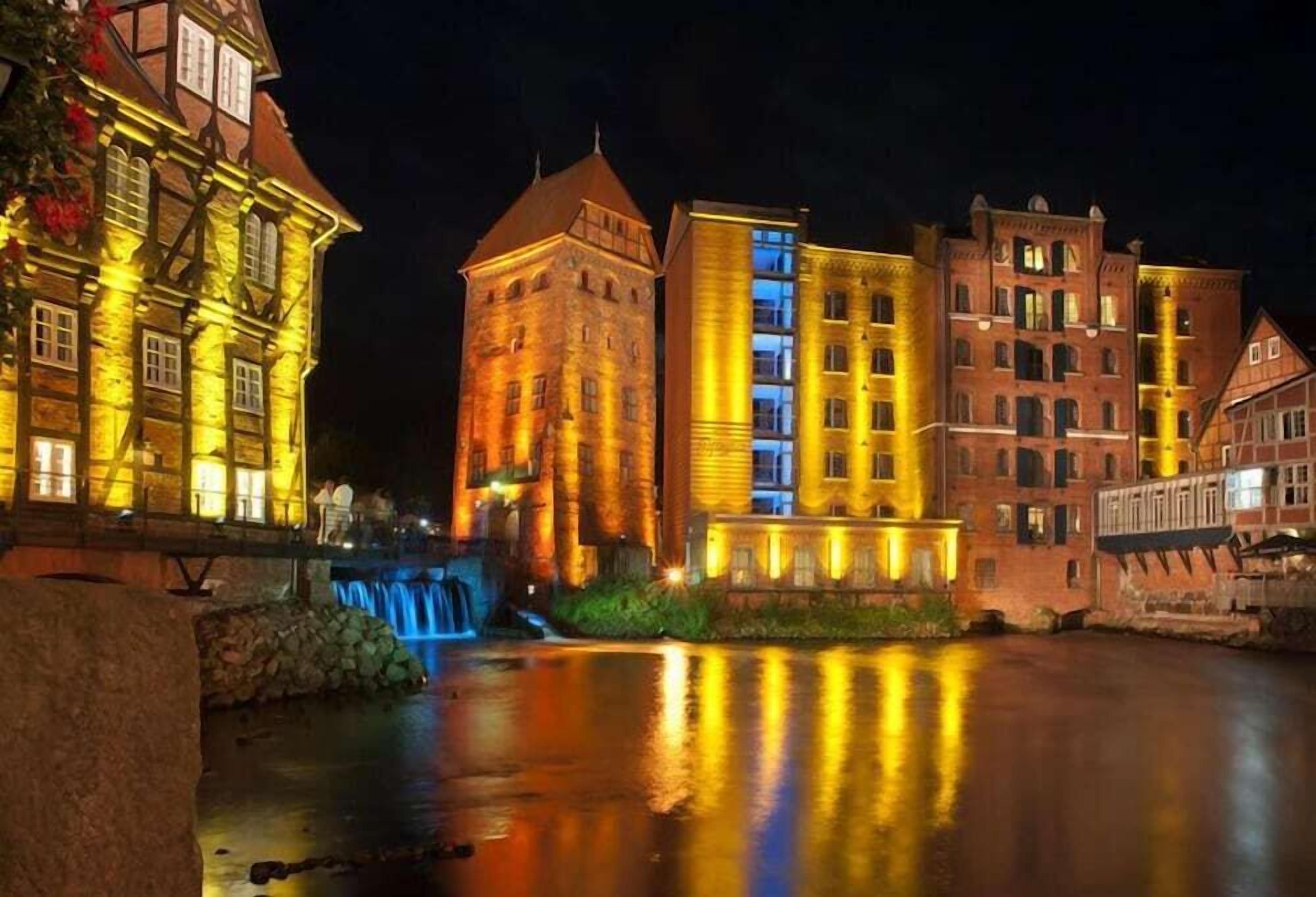 Ein historisches Gebäude mit Turm, beleuchtet durch warmes Licht, neben einem modernen Bau, beide Spiegelungen auf einer ruhigen Wasseroberfläche.