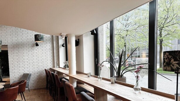 Een moderne bar met een lange houten toonbank, verschillende barkrukken en grote ramen met uitzicht op de straat.