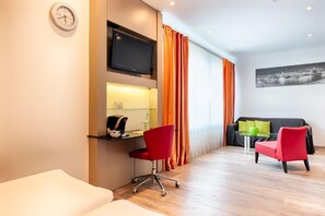 Ein modernes Hotelzimmer mit Schreibtisch, zwei roten Sesseln, einer Couch und einem Flachbildfernseher.