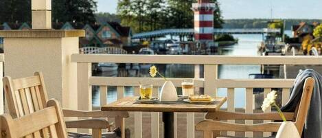 A wooden deck with a table and chairs, overlooking a body of water with a lighthouse and buildings in the background.