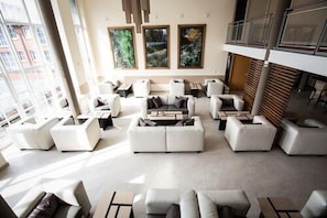 Ampla área de estar com diversas opções de assento, incluindo sofás e poltronas, dispostas ao redor de uma mesa de centro. Janelas grandes proporcionam luz natural e vista para o exterior.