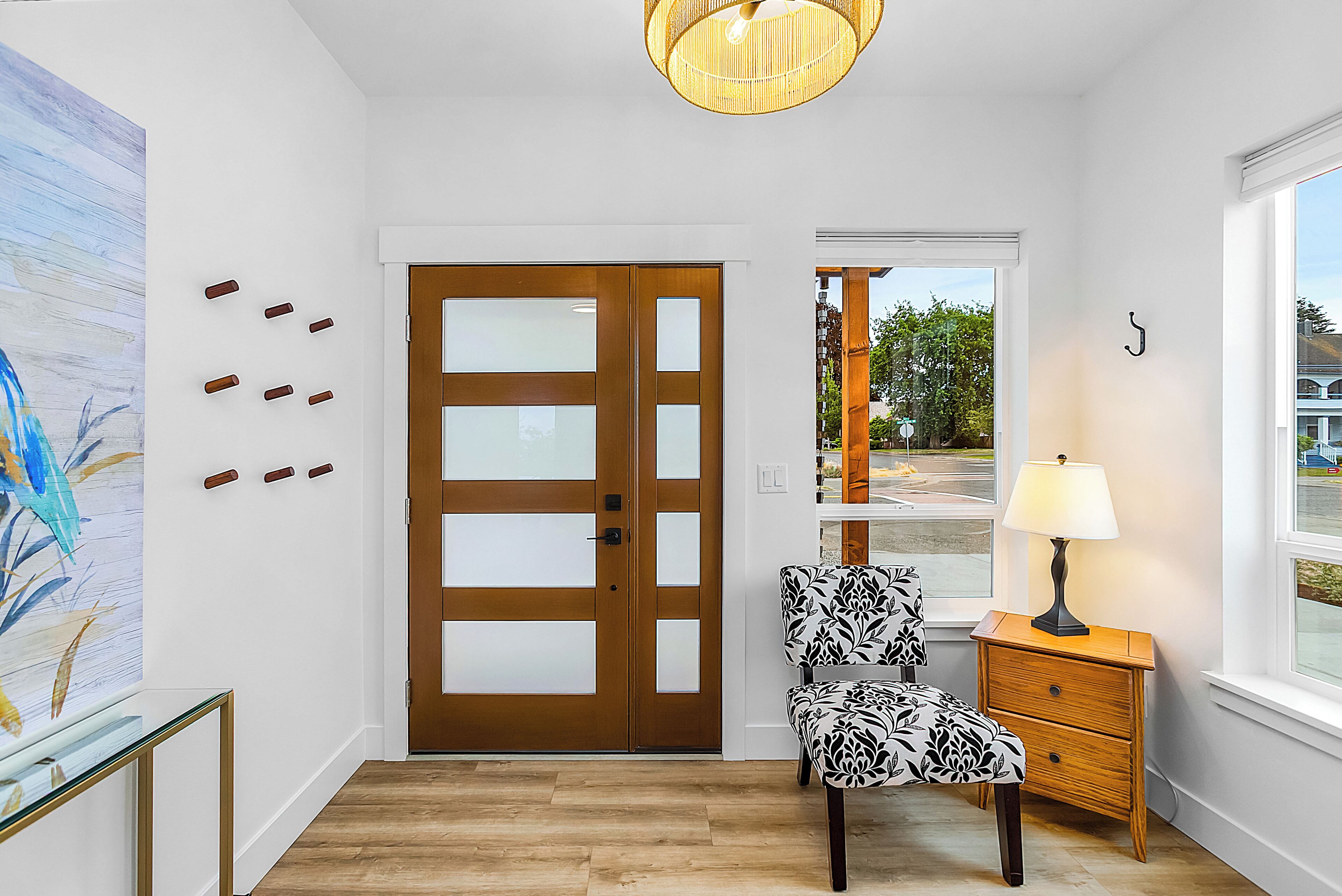 A room with a wooden door, a patterned chair, a small wooden nightstand with a lamp, and a large window with a view of the outside.