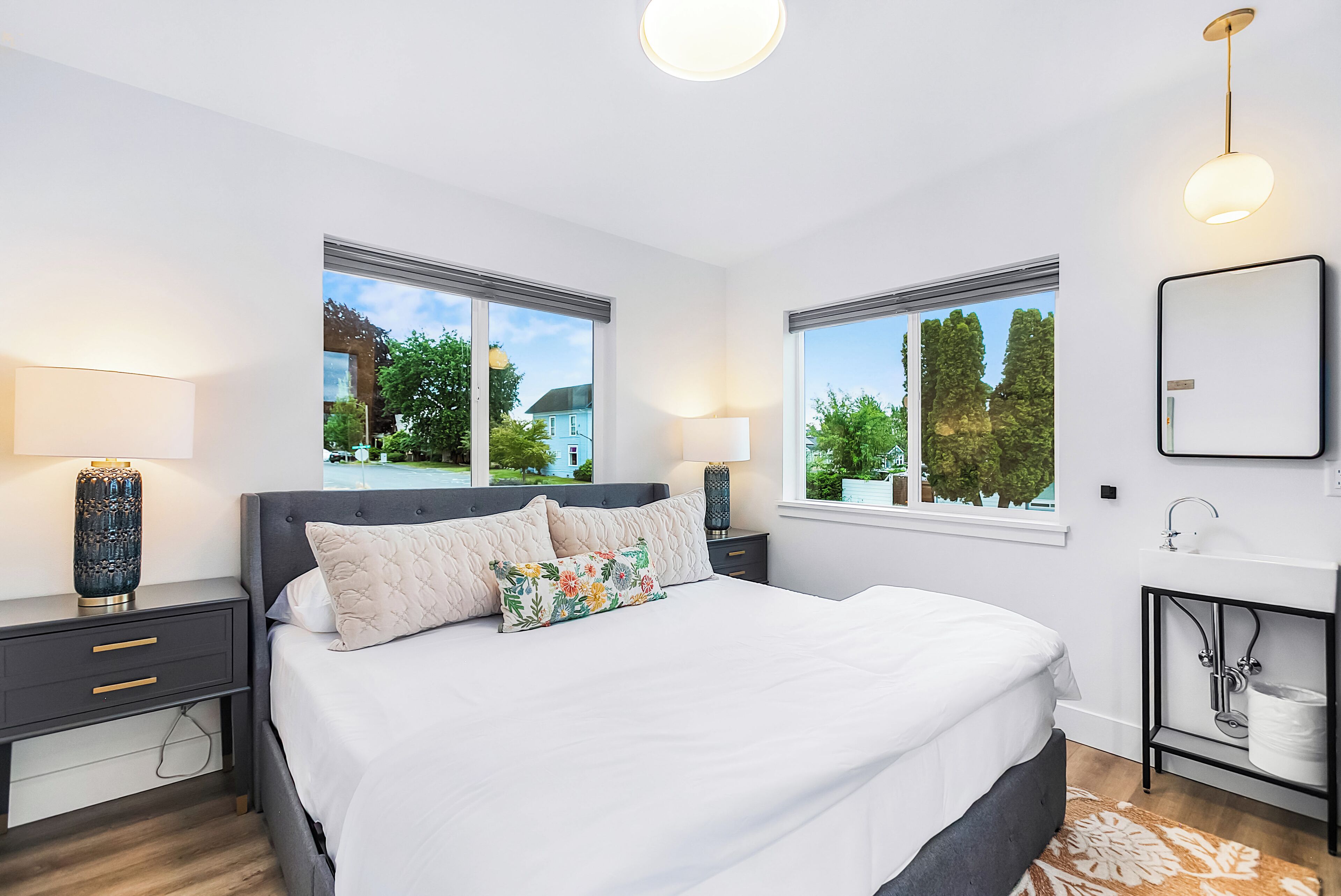 A modern bedroom with a large bed, two bedside tables, a nightstand, a bedside lamp, a wall-mounted mirror, and a window with a view of greenery and a building.