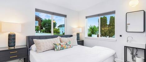 A modern bedroom with a large bed, two bedside tables, a nightstand, a bedside lamp, a wall-mounted mirror, and a window with a view of greenery and a building.