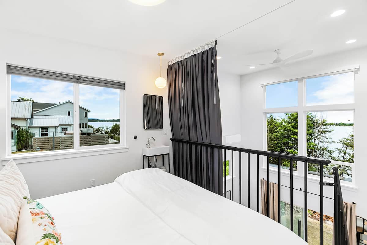 A bedroom with a large bed, a window with a view of a house and trees, a small bedside table with a lamp, and a balcony with a railing.
