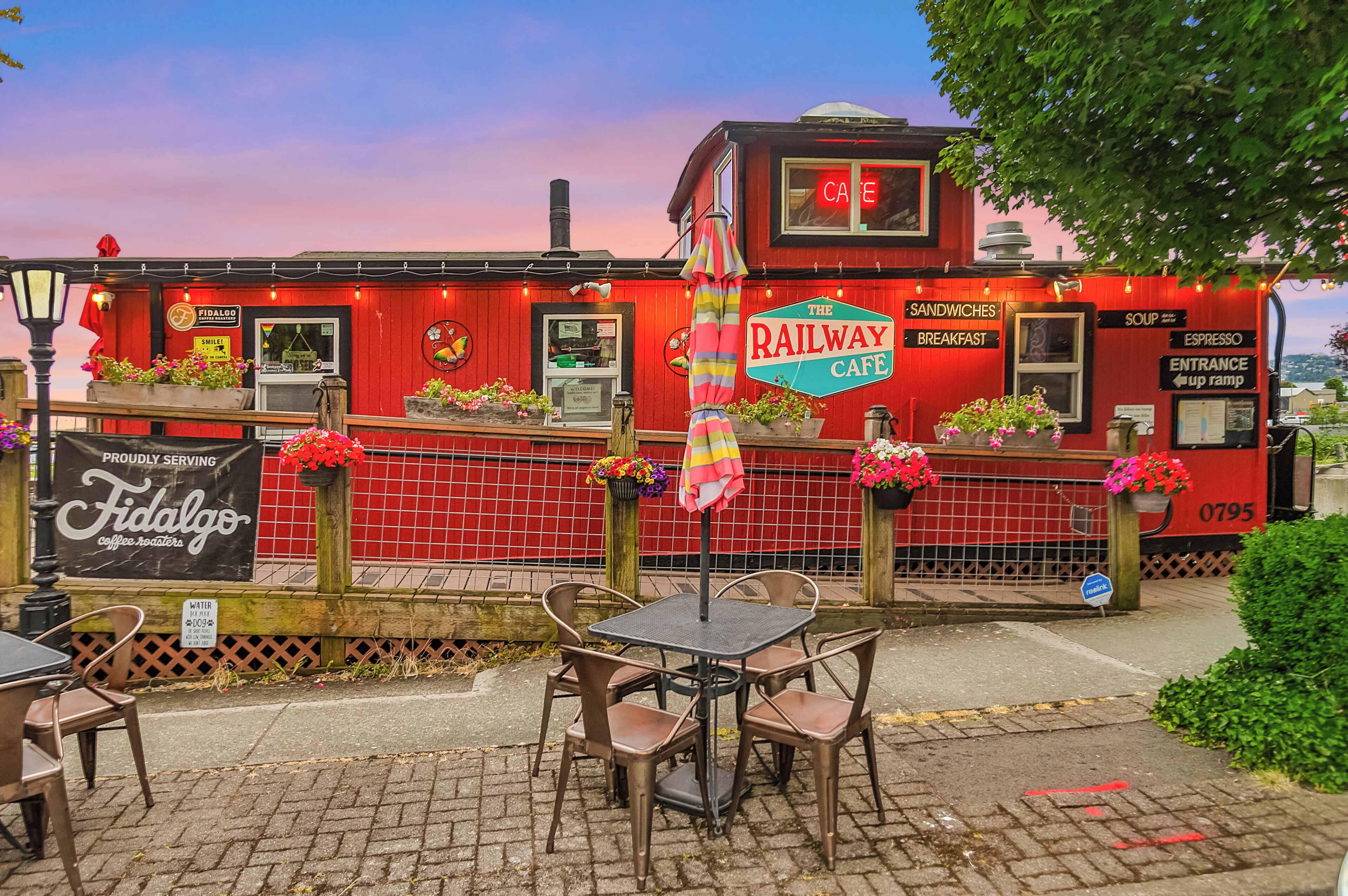 Un bâtiment rouge avec une enseigne indiquant « The Railway Cafe » et un panneau lumineux au-dessus de l’entrée. Des tables et des chaises sont disposées à l’extérieur, devant le bâtiment.
