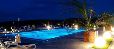 A swimming pool with lounge chairs and a statue, surrounded by palm trees and well-lit pathways.