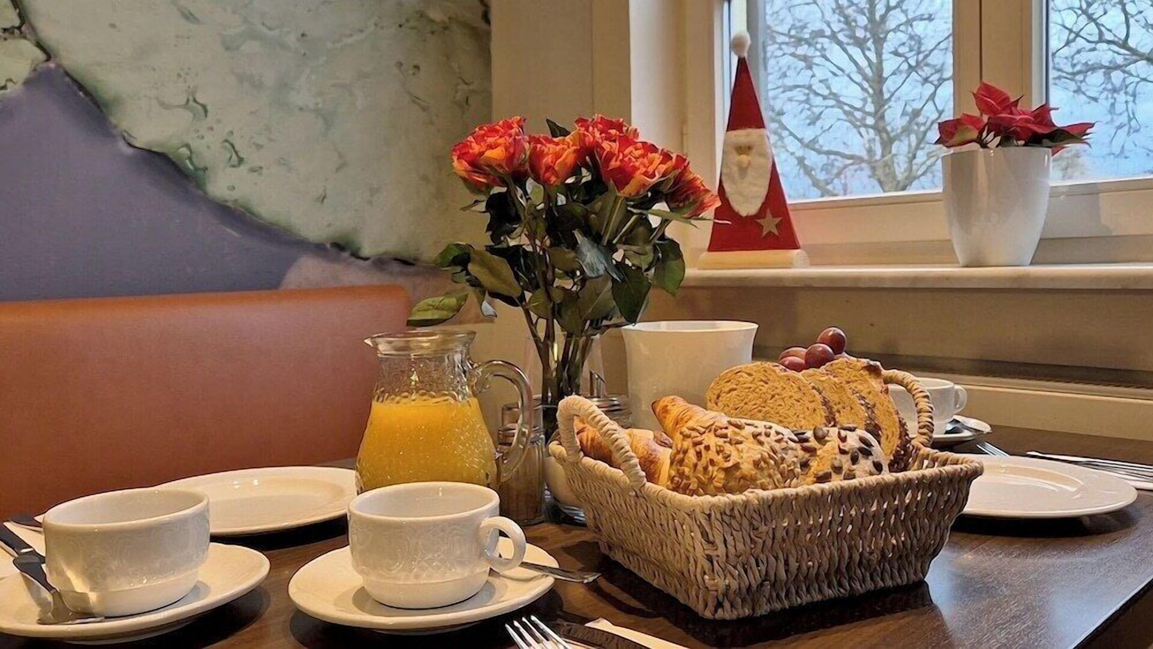 Ein Frühstückstisch mit einem Korbwannen-Arrangement von Croissants, einem Krug Orangensaft und einem Strauß roter Blumen.
