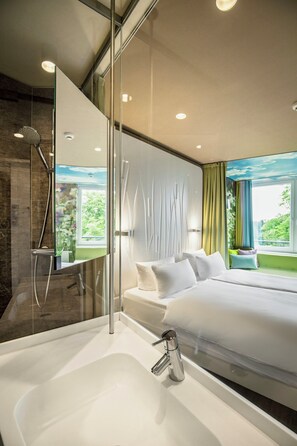 A modern hotel room with a glass shower enclosure, a large bed with white linens, and a view of greenery through the window. - A-ja Bad Saarow. Das Resort (Bad Saarow)