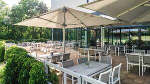 An outdoor patio with tables and chairs, large umbrellas, and a glass building in the background. - A-ja Bad Saarow. Das Resort (Bad Saarow)