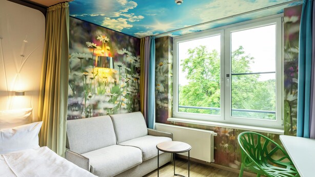 A room with a sofa, a small round table, a green chair, a window with a view of greenery, and a ceiling mural. - A-ja Bad Saarow. Das Resort (Bad Saarow)