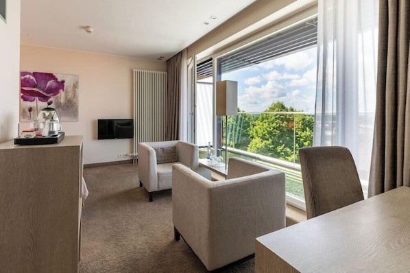 A hotel room with a large window, a flat-screen TV, a sofa, a table, and a decorative item on the desk. - Thomas Hotel Spa Lifestyle (Husum)